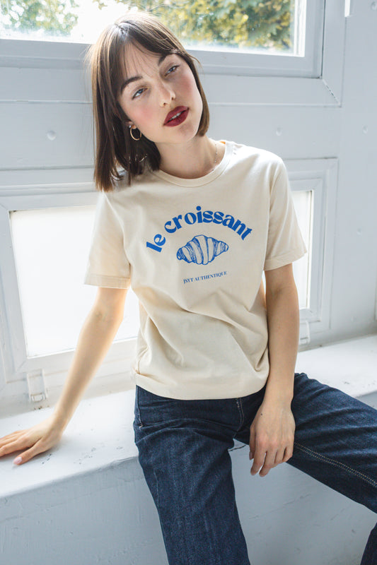 Woman wearing a beige t-shirt with blue text and graphics, sitting on a windowsill. Le Croissant t-shirt.
