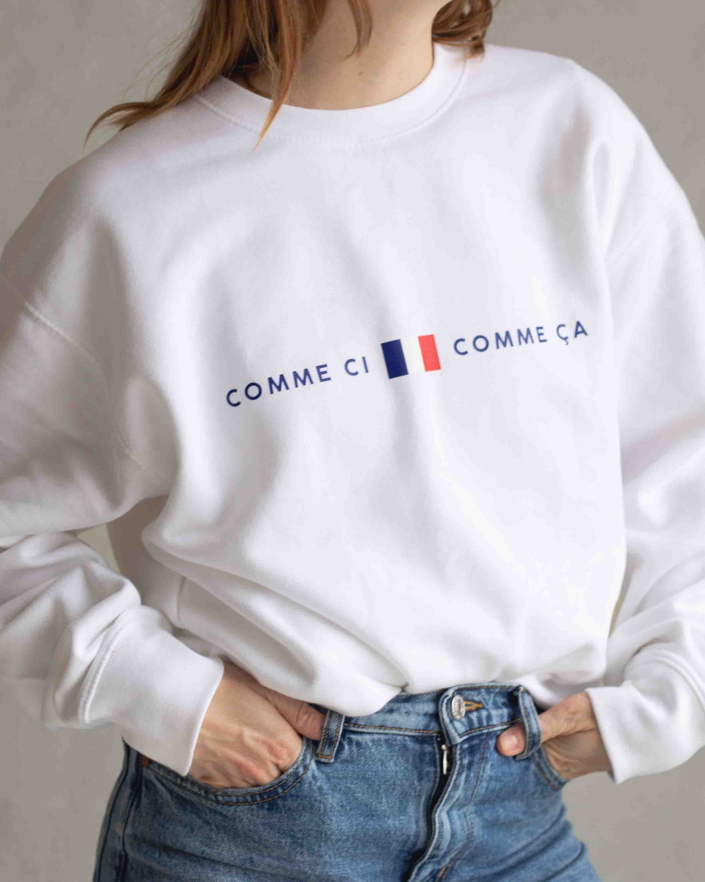 White sweatshirt with 'Comme ci comme ca' text and french flag worn by a person against a neutral background
