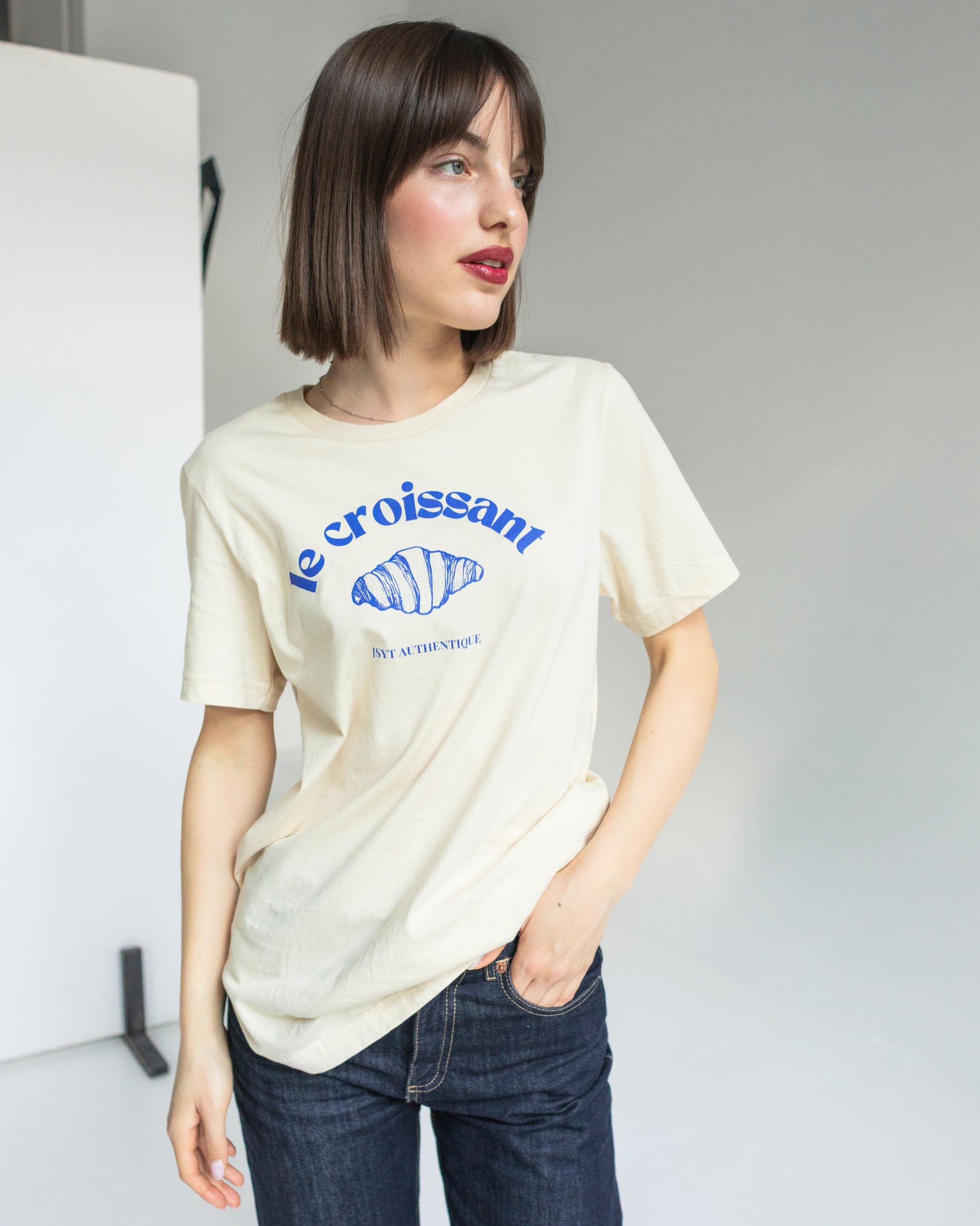 Woman wearing a beige t-shirt with blue text and graphics against a plain background. Le croissant tee in beige colour. 