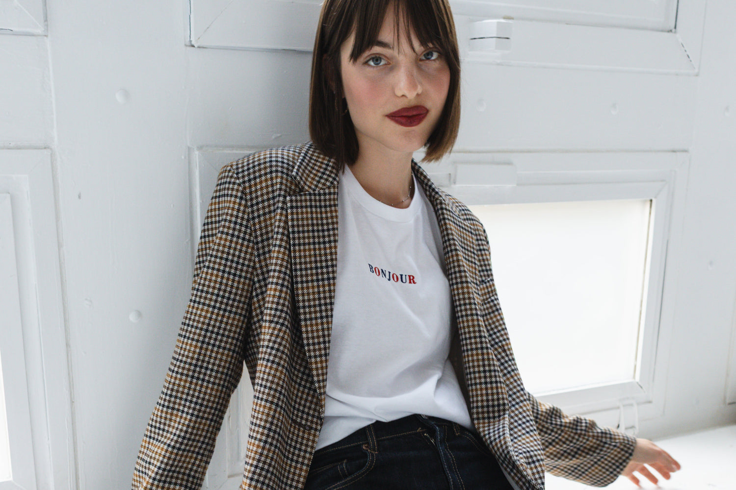 Woman sitting on a windowsill wearing a checkered blazer and white t-shirt.