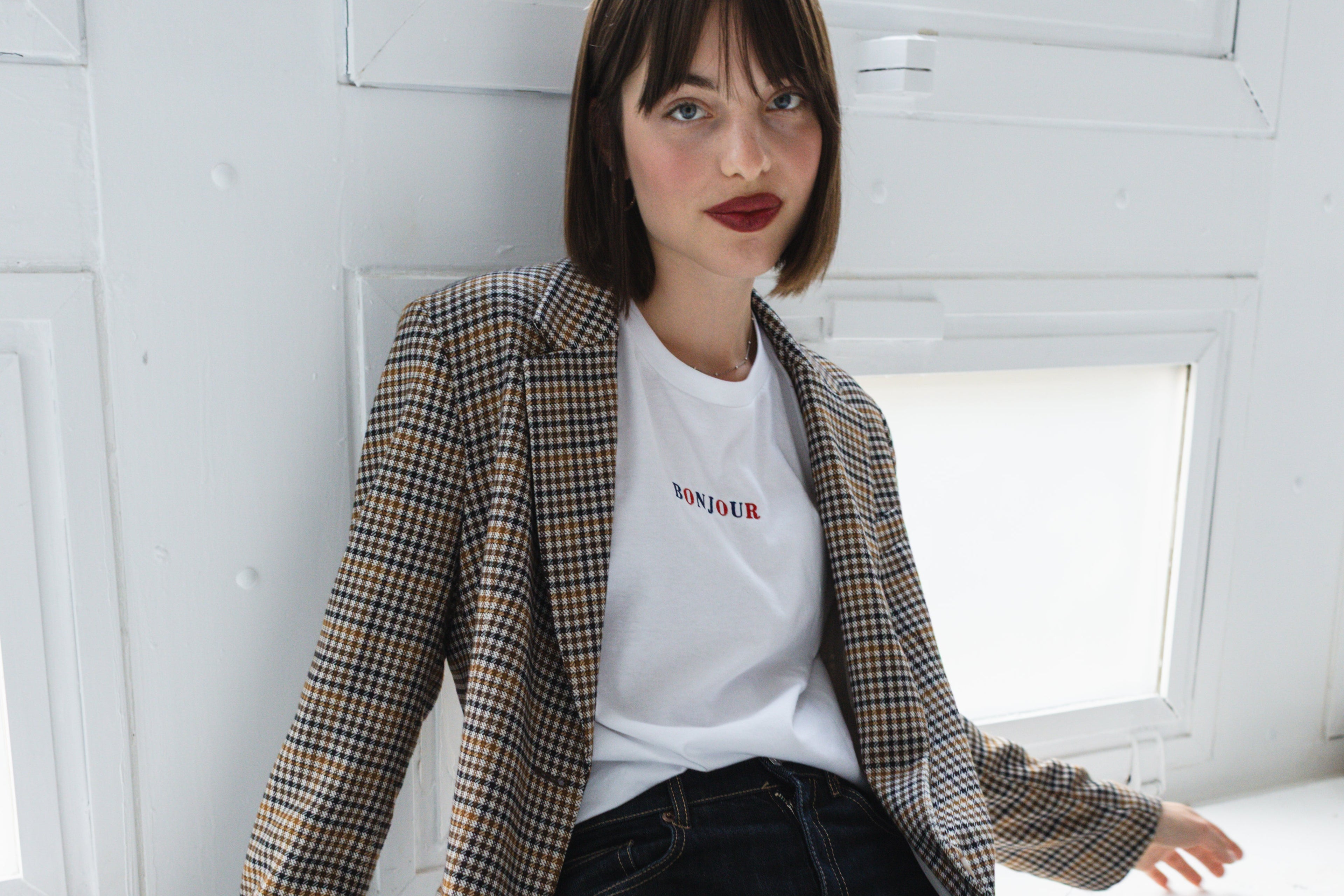 Woman sitting on a windowsill wearing a checkered blazer and white t-shirt.