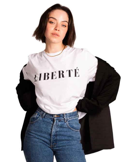 Person wearing a white sweatshirt with 'LIBERTé' text, blue jeans, and a black coat on a light background